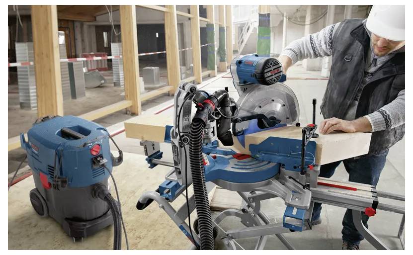A person operates a miter saw on a construction site, cutting a wooden beam. Safety equipment and a dust extractor are in use.
