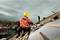 A construction worker wearing a yellow hard hat is sawing wood with an electric saw on a roof. A city view can be seen in the background.