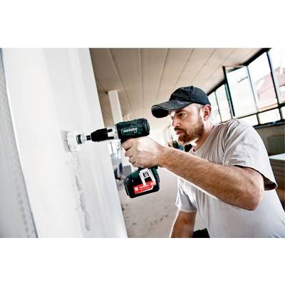 A man is using a drill in an unfinished room. He is wearing a baseball cap and is focused on working on the wall.