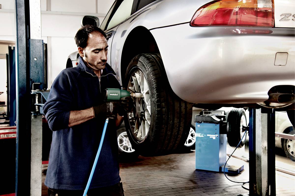 A mechanic is changing the tyre of a raised vehicle in a workshop. He is using an electric tool.