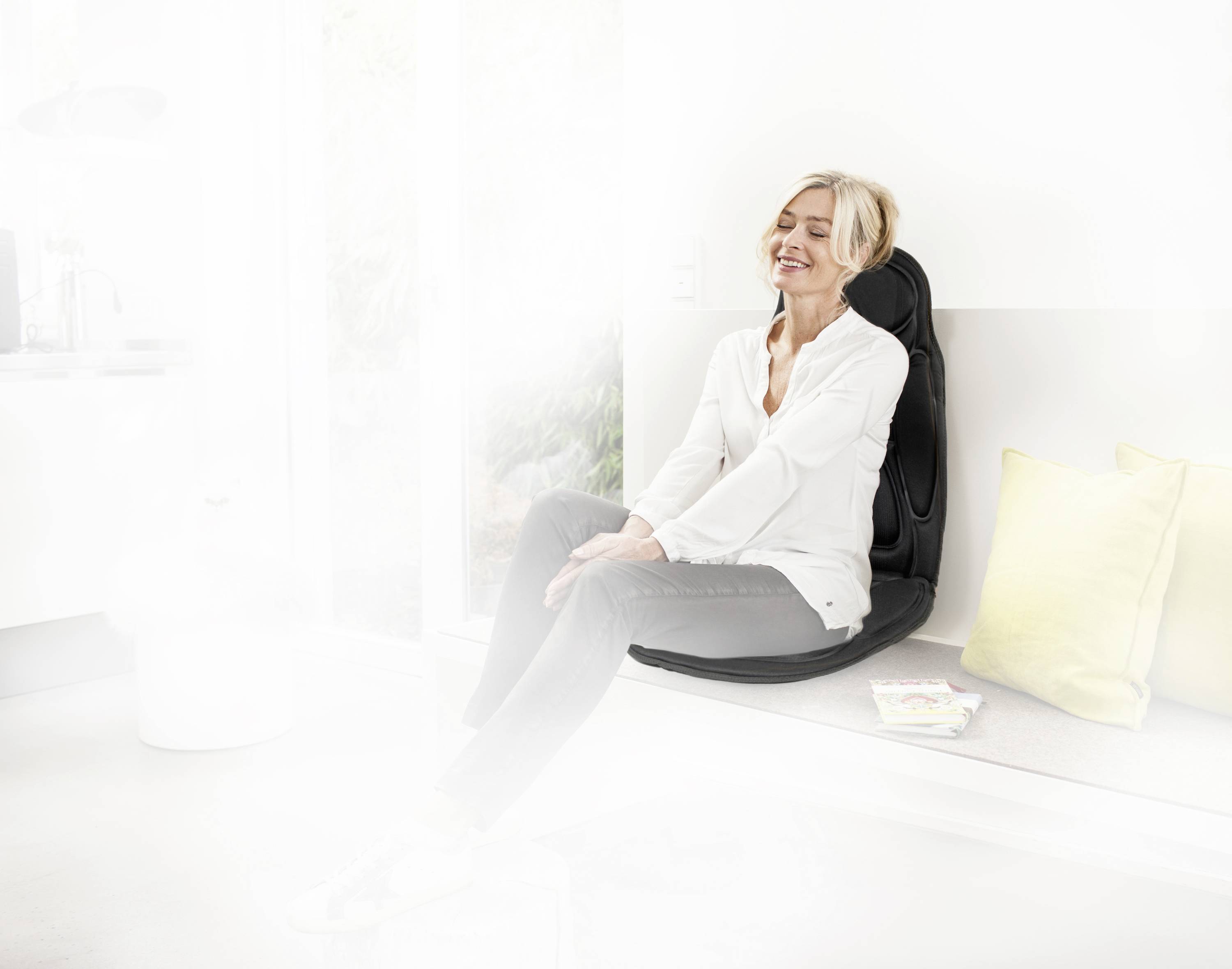 A woman is sitting smiling on a massage seat cushion in a bright room. Two pillows and a magazine are lying beside her.