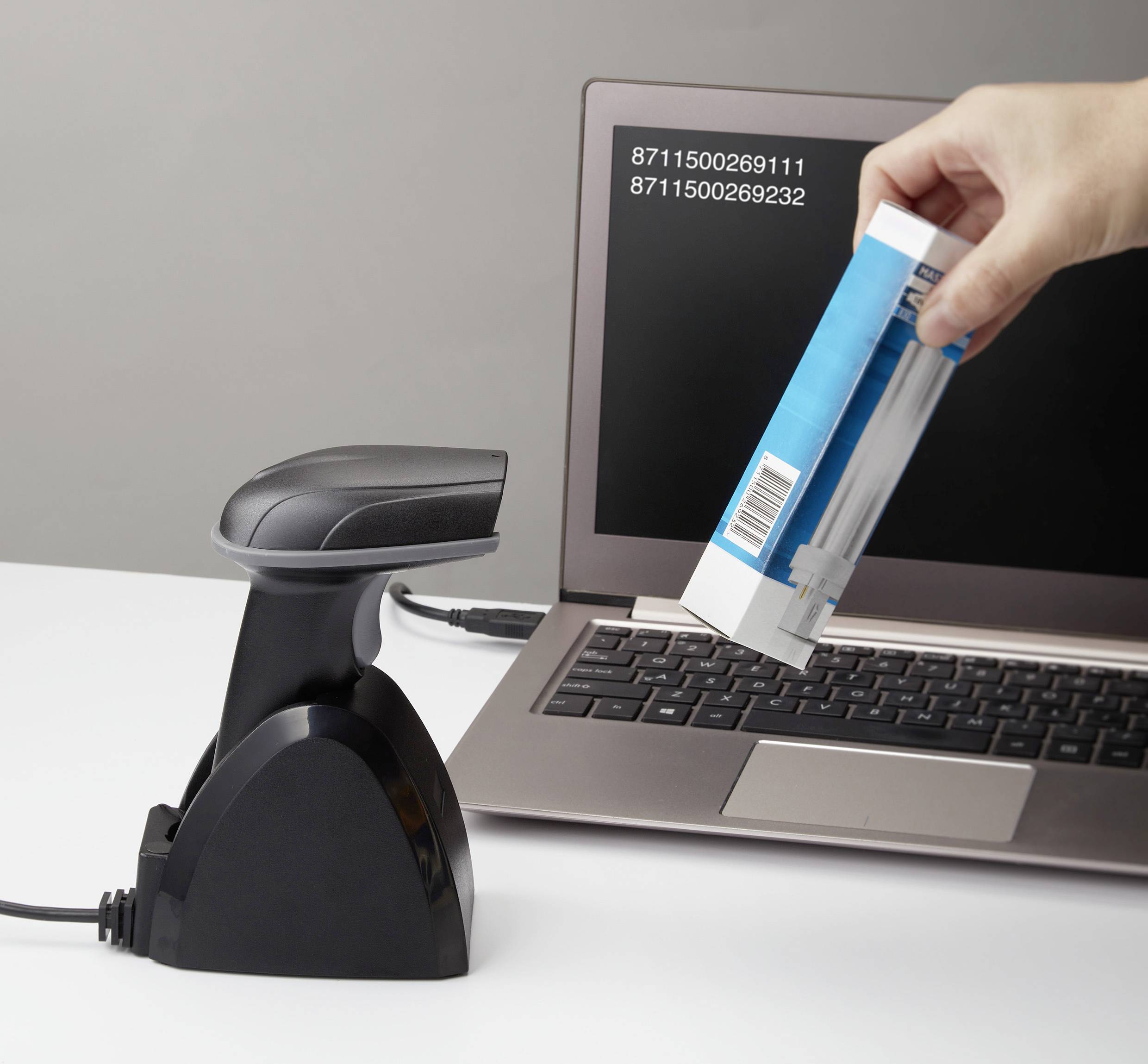 A hand is scanning a barcode on a package using a scanner. In the background, a laptop displays the scanned numbers.