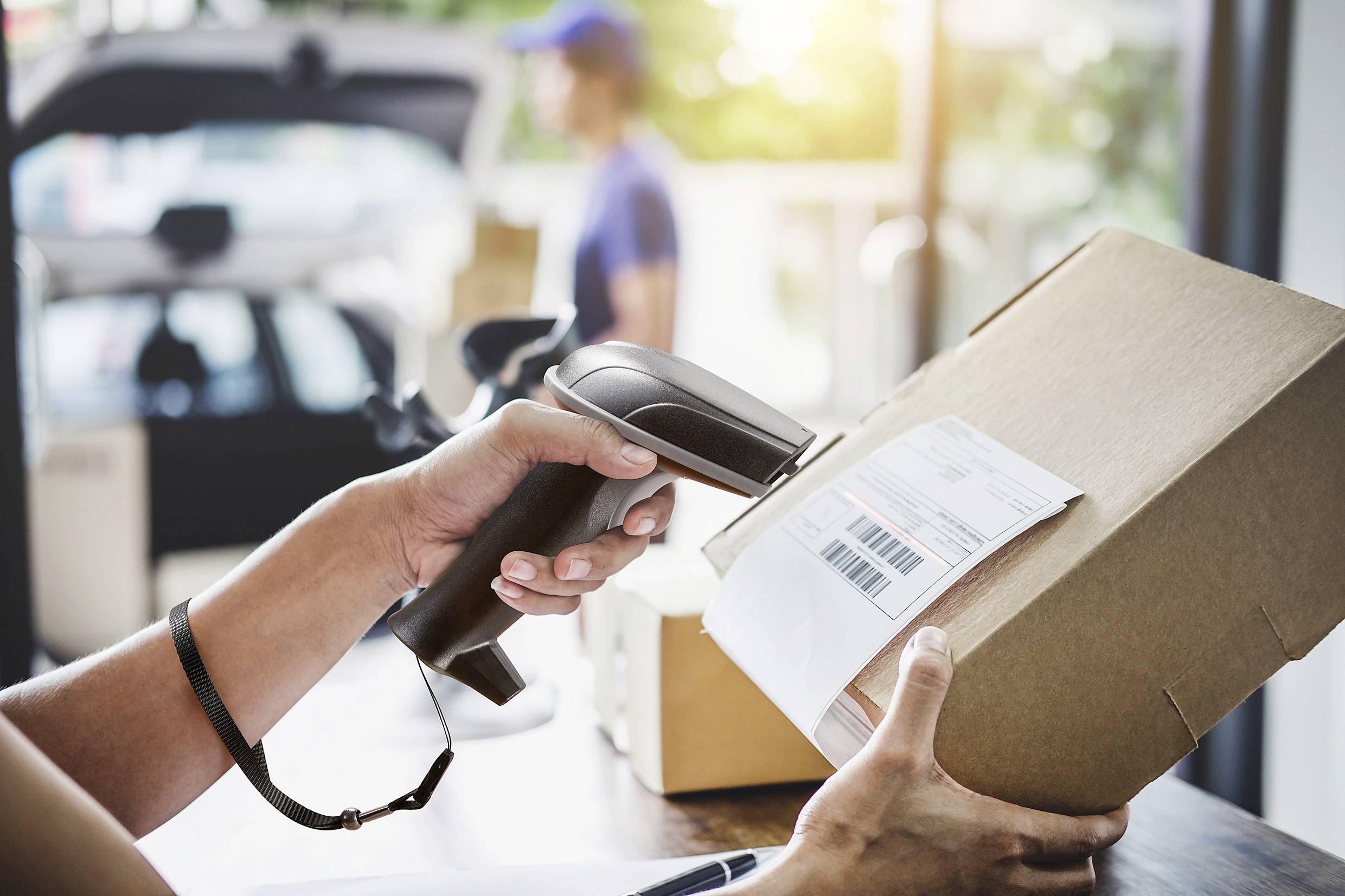 A person is scanning a package with a barcode scanner. In the blurred background, a second person and a vehicle can be seen.