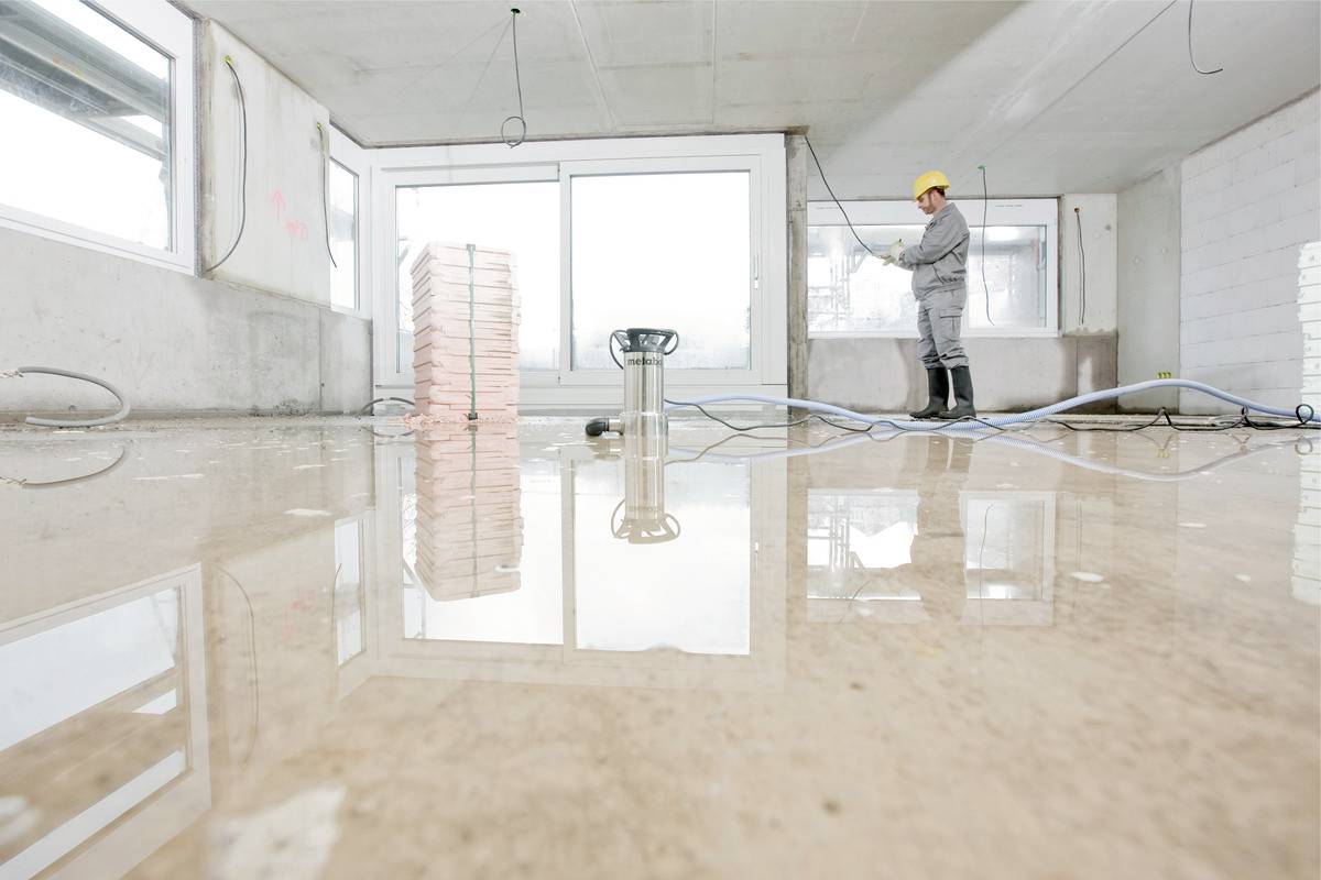 A construction worker in protective clothing and a hard hat is inspecting a construction site with large windows. The floor is wet and reflective.