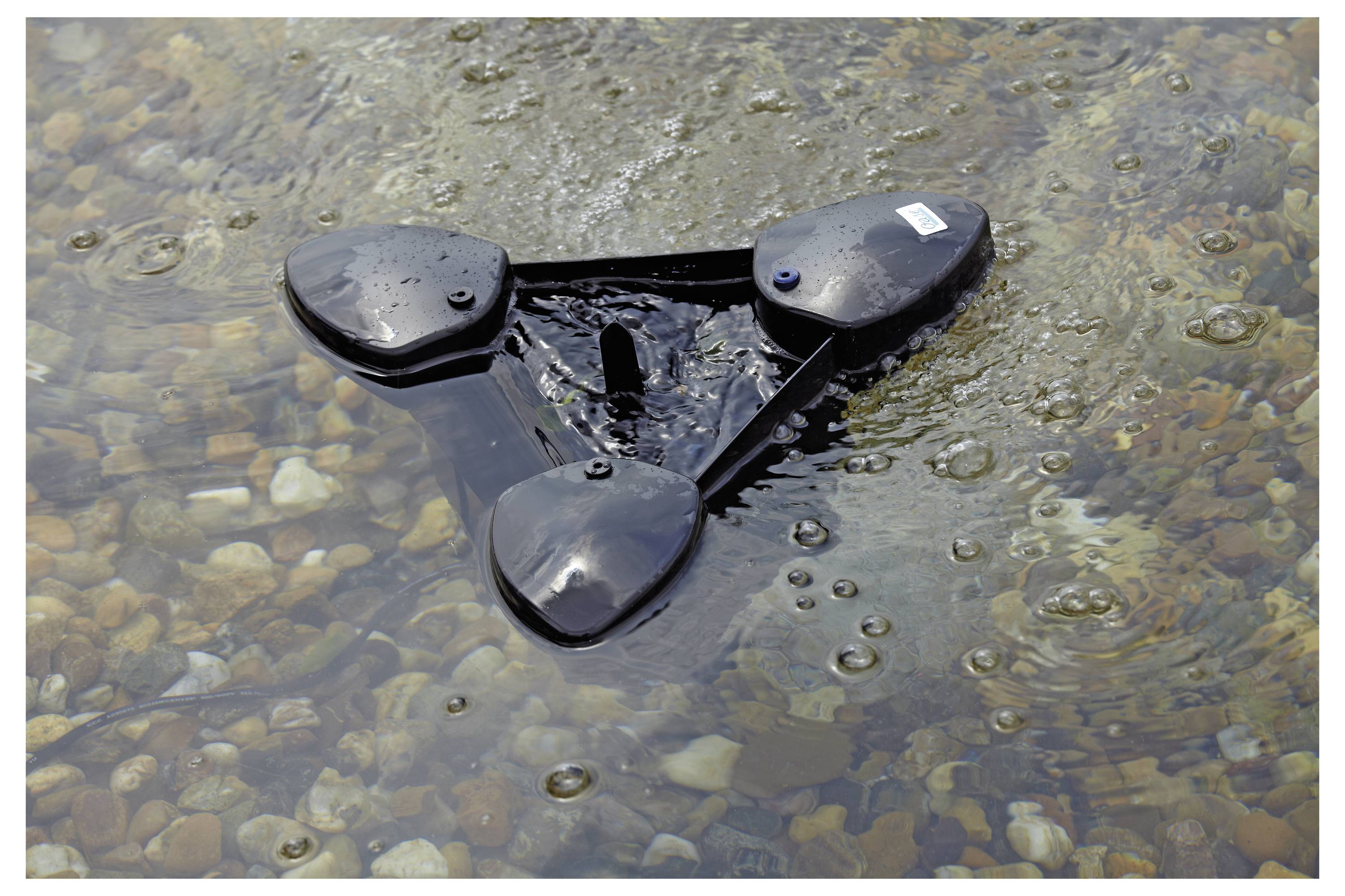 A black, three-paddle debris removal device floats on a rocky water surface, collecting litter in a stream or pond.