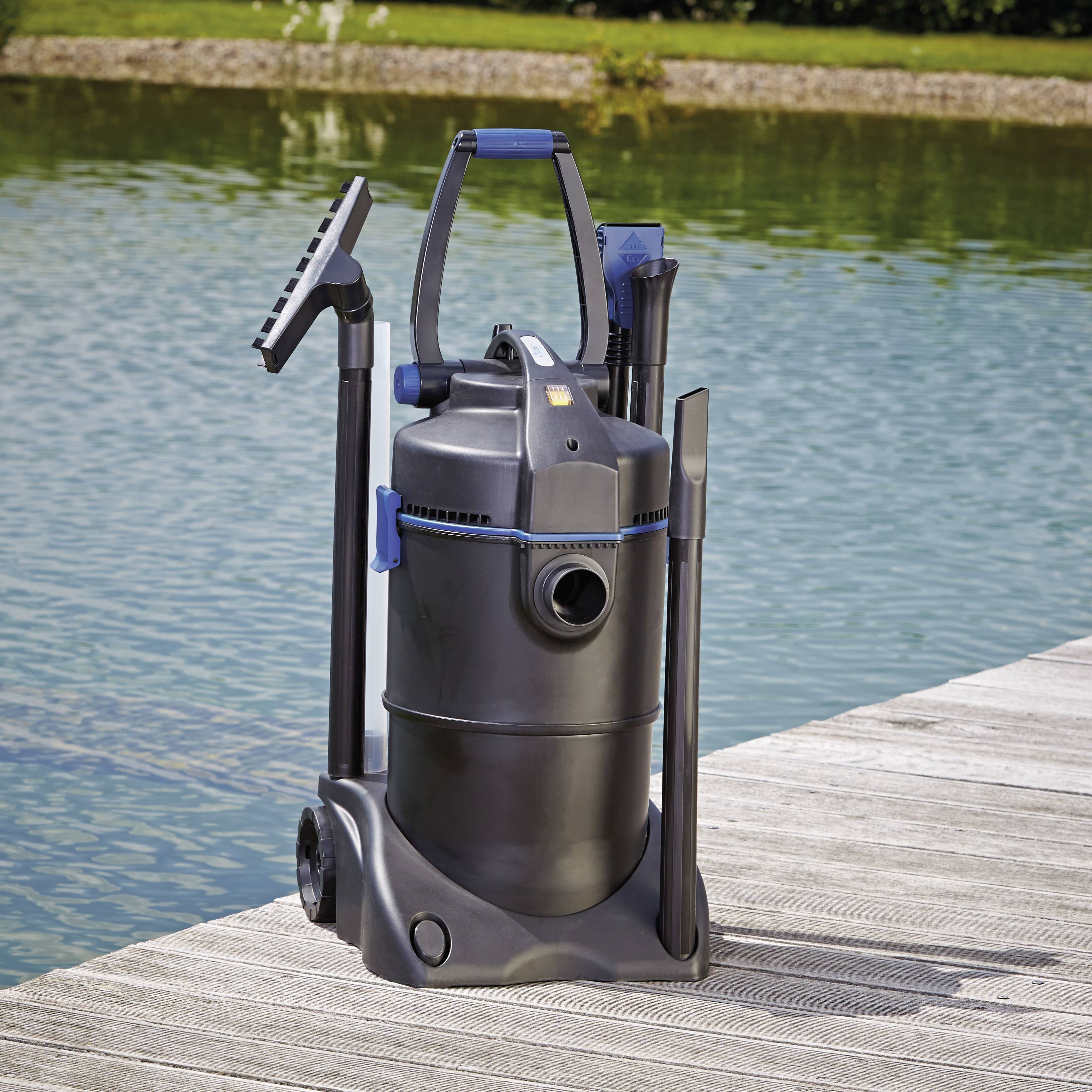 A black pond vacuum cleaner with attachments stands on a wooden deck by a pond. The pond and greenery are visible in the background.