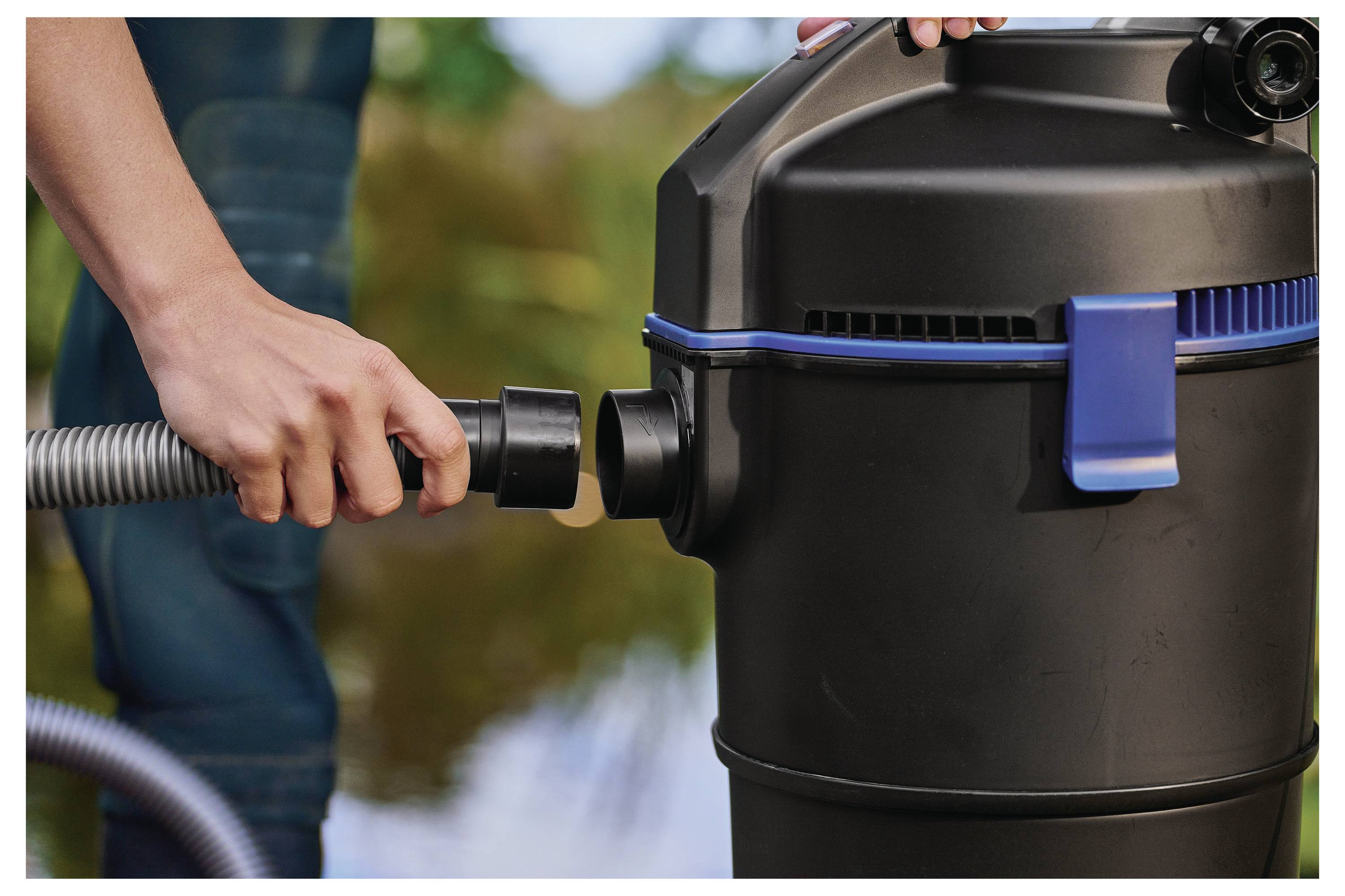 A person connects a hose to a black outdoor vacuum with a blue latch, near water.