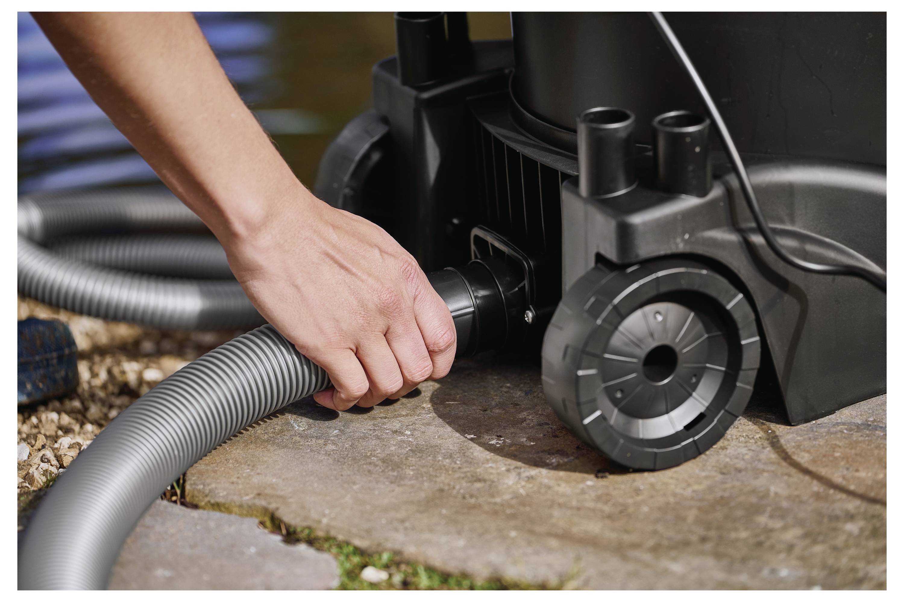 A hand connects a flexible hose to a black outdoor vacuum device on a stone surface, with a pond visible in the background.