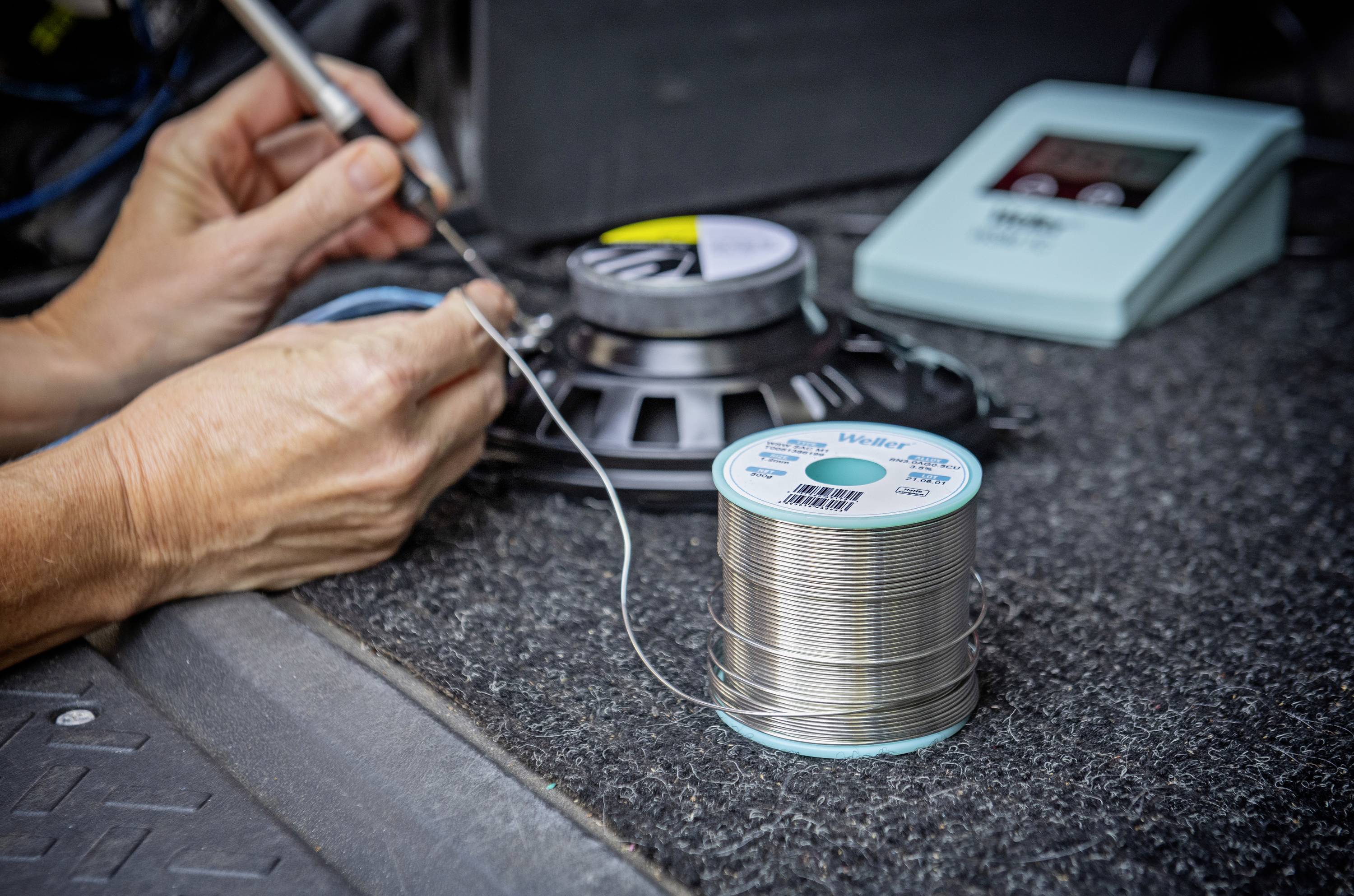 A person is soldering wires on a speaker, with a reel of solder in the foreground. Electronic work is being carried out.