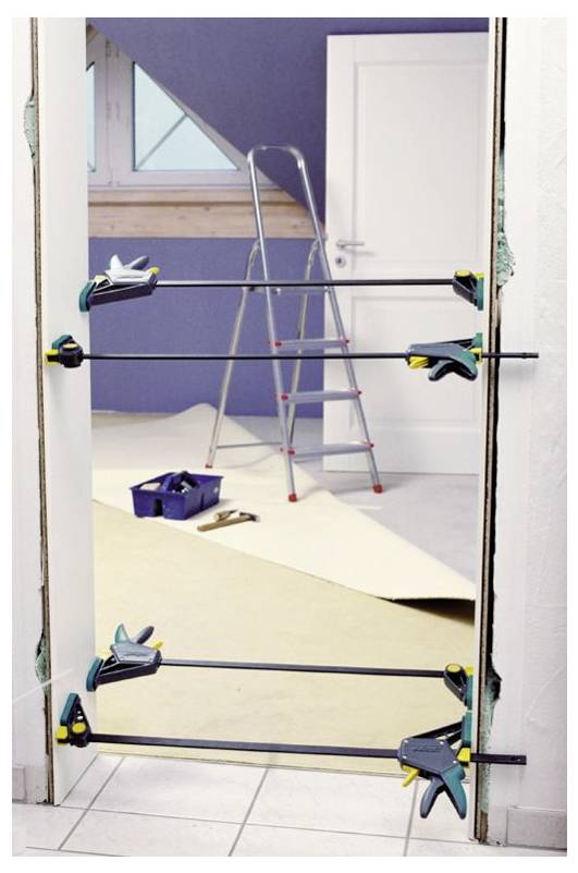 A door frame held in place by clamps during installation or repair. A stepladder and toolbox are in the background, indicating ongoing construction work.