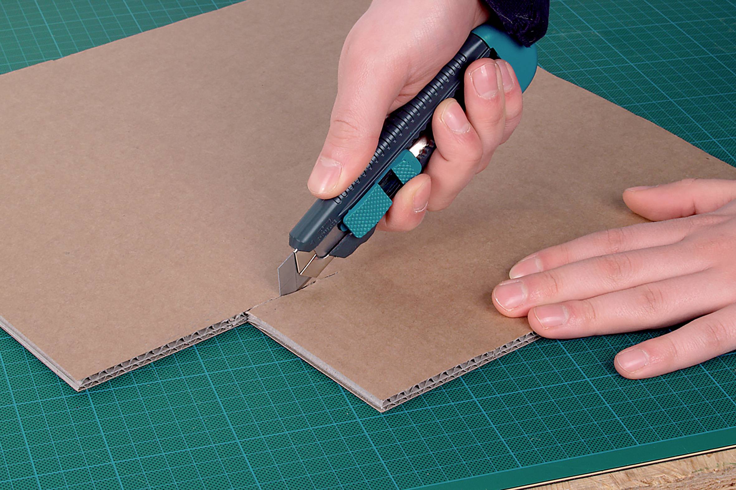 A hand is cutting cardboard with a utility knife on a green cutting mat.