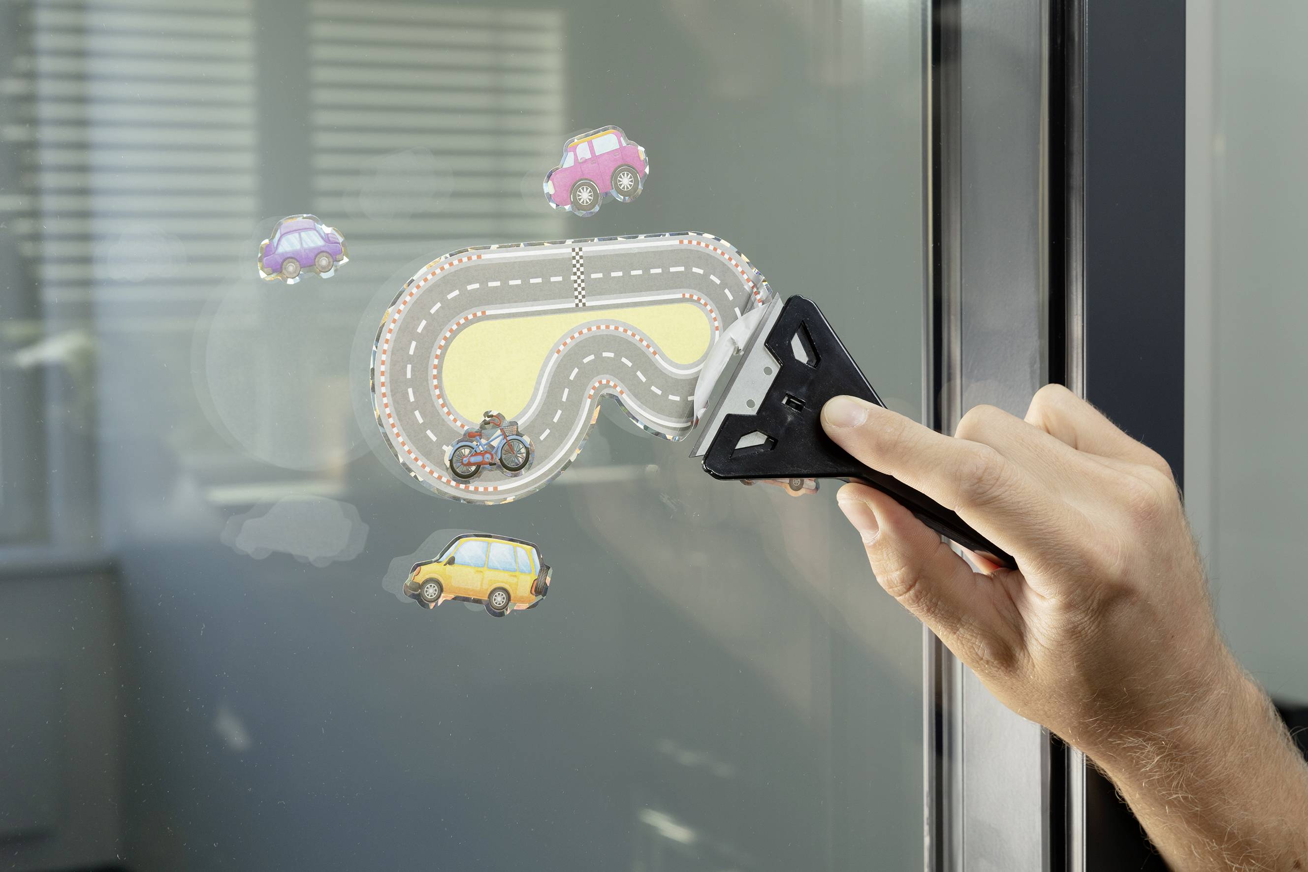 A hand holds an ice scraper against a windscreen, removing a sticker with a road and vehicles. Background blurred.
