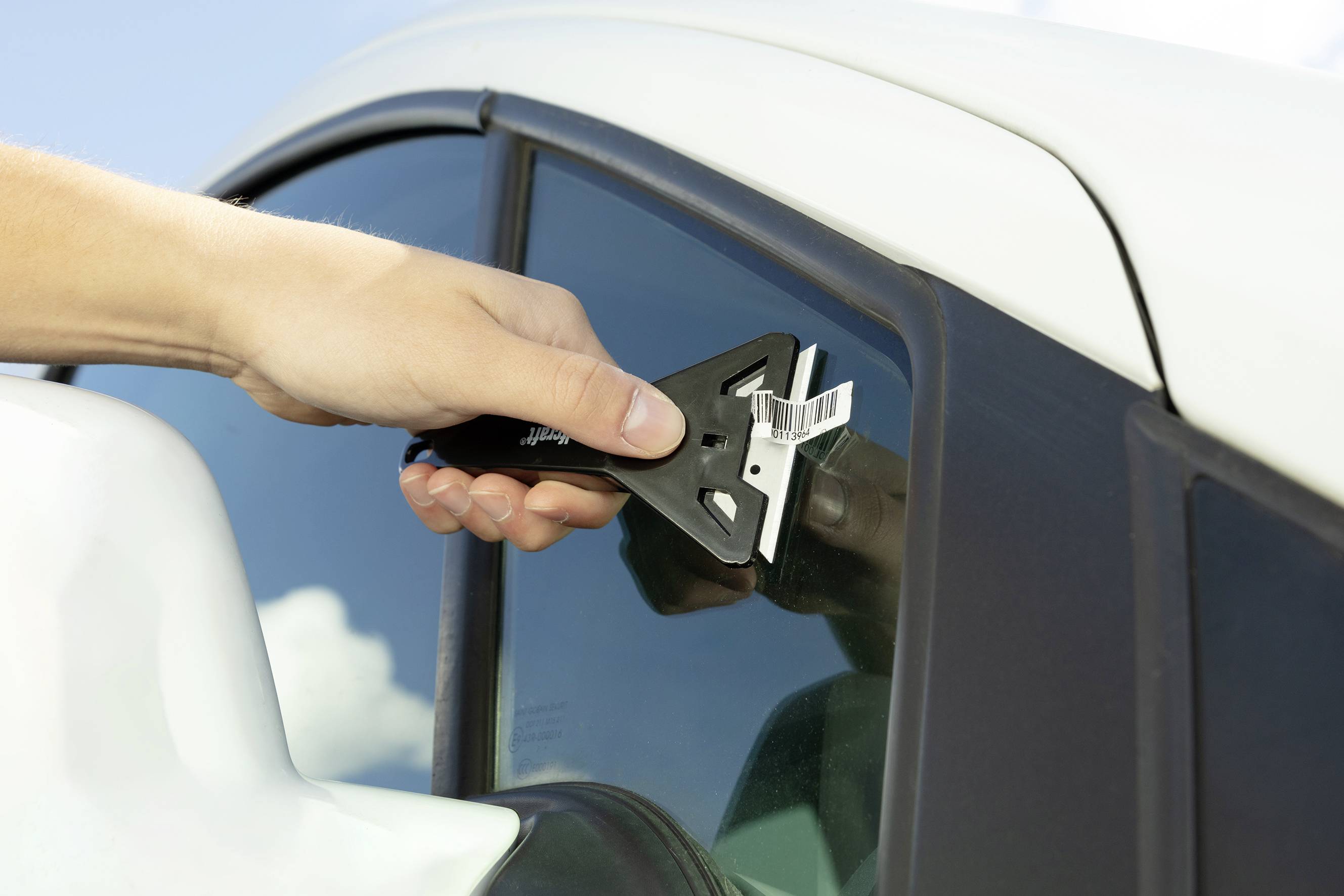 A hand holds an ice scraper and removes ice from a car windscreen. The car is white, and it is a sunny day.