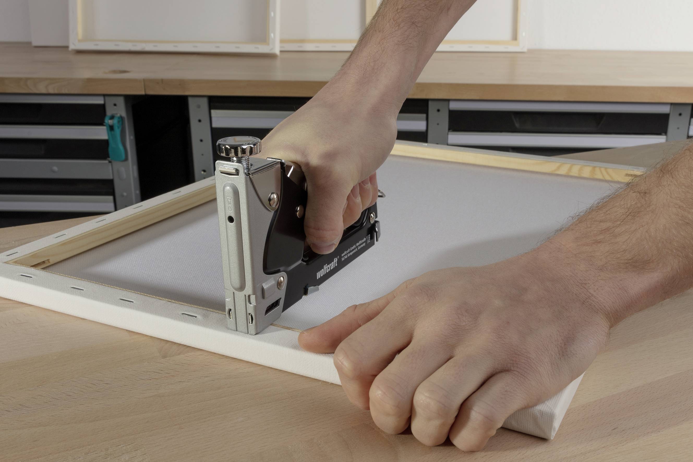 A person is attaching a canvas to a wooden frame using a staple gun, on a workbench in a workshop setting.