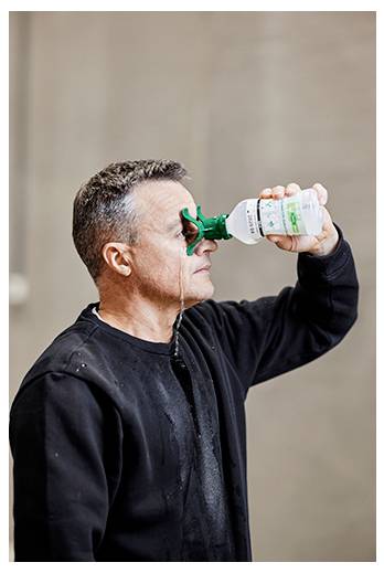 A man in a black shirt uses a portable eyewash bottle to rinse his eye, standing against a neutral background.