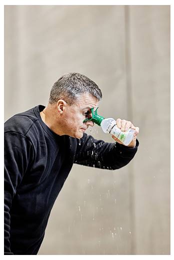 A person in a black shirt uses an eye wash bottle to cleanse their eyes, tilting their head slightly forward as liquid flows down.