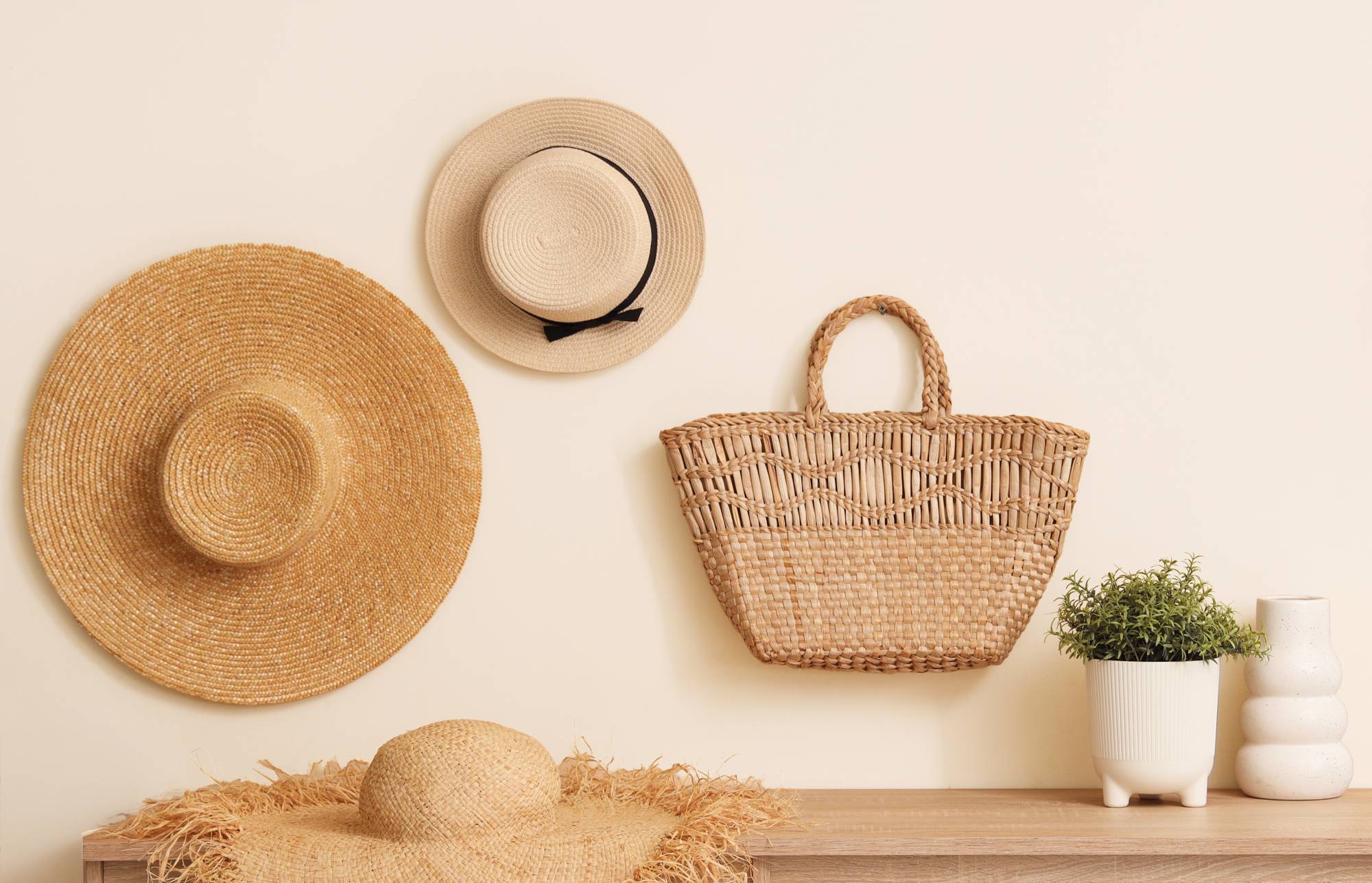Three woven hats and a wicker basket hang above a wooden shelf with a straw hat, potted plant, and white vase against a beige wall.