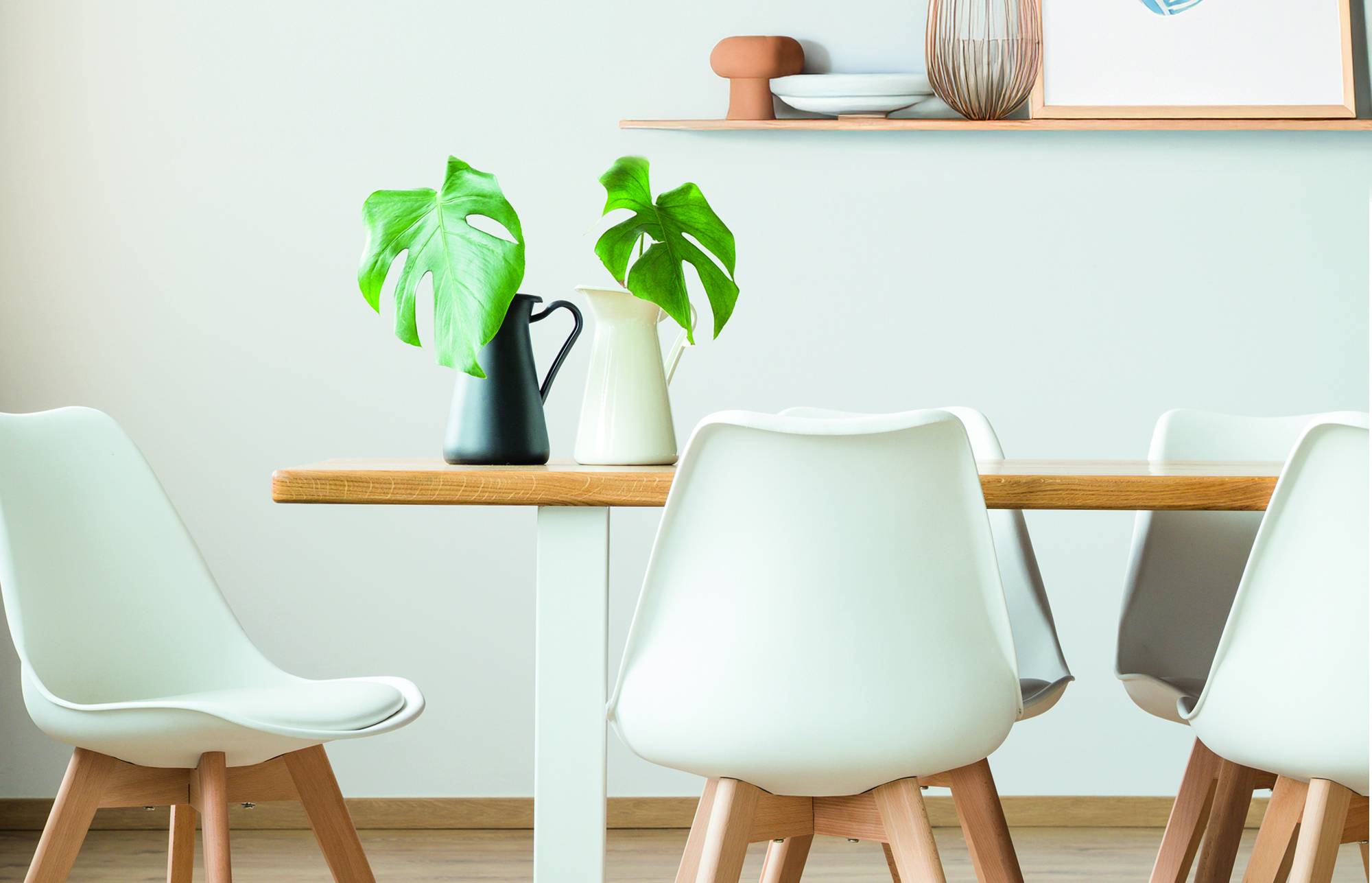 Modern dining area with white chairs around a wooden table, two green plants in vases, and wall shelves with decor.