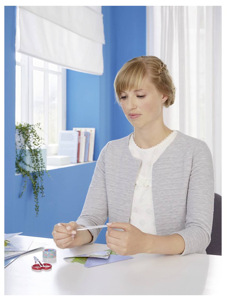 A woman sits at a table holding a piece of thread, with crafting materials nearby in a bright, blue-walled room.