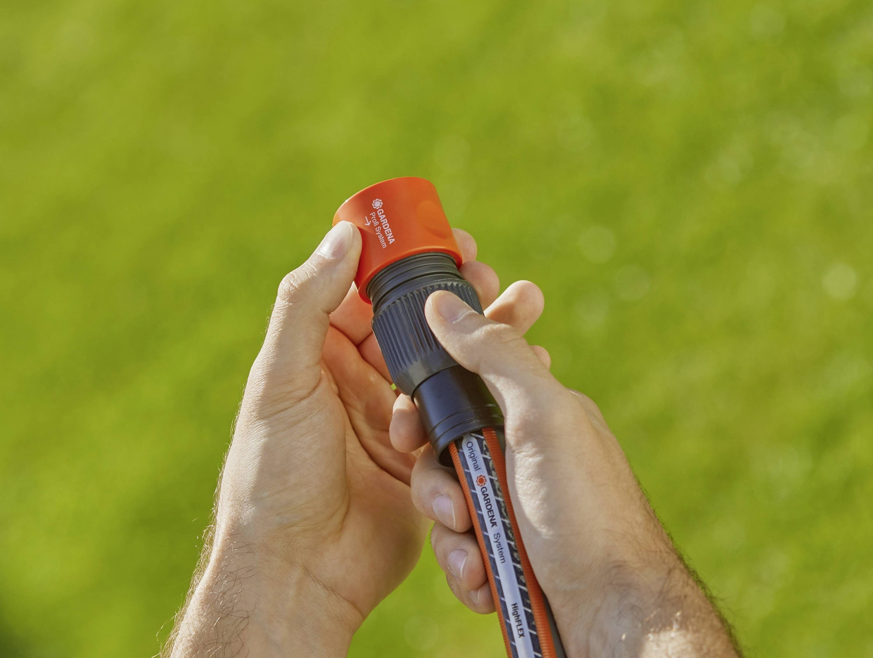 A person is holding a garden hose connector with a red end in their hand, apparently preparing it for use. Grass is visible in the background.