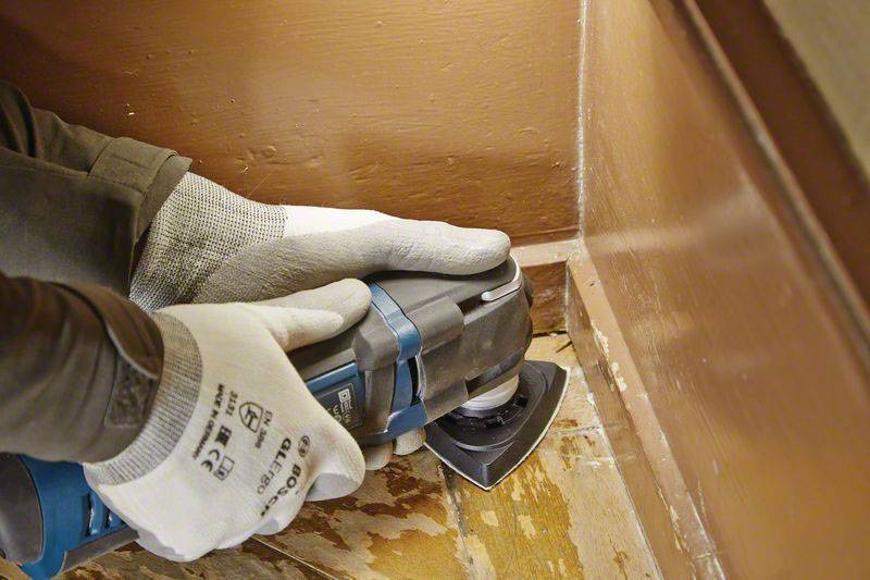 A worker is sanding the floor in the corner of a room using an electric multi-tool. He is wearing gloves for protection.