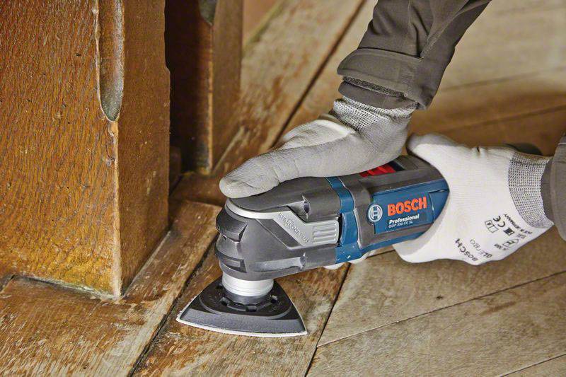 A person is sanding a wooden corner with a Bosch multi-tool. The scene depicts precise craftsmanship on a wooden floor.