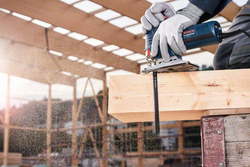 A person is cutting wood with an electric jigsaw on a construction site. Sunlight is shining through wooden beams in the background.