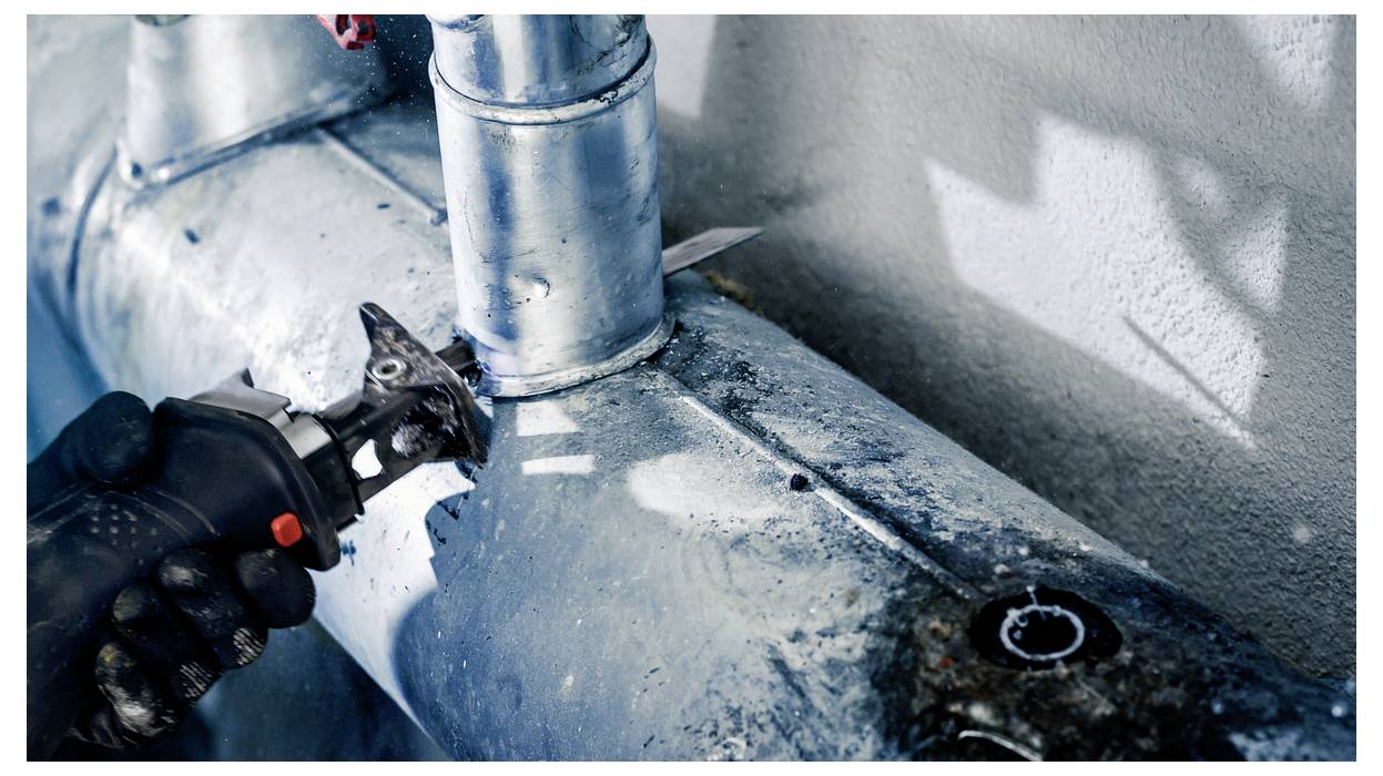 A person uses a power tool to cut a metal pipe, with sparks visible. The scene suggests industrial or plumbing work in progress.