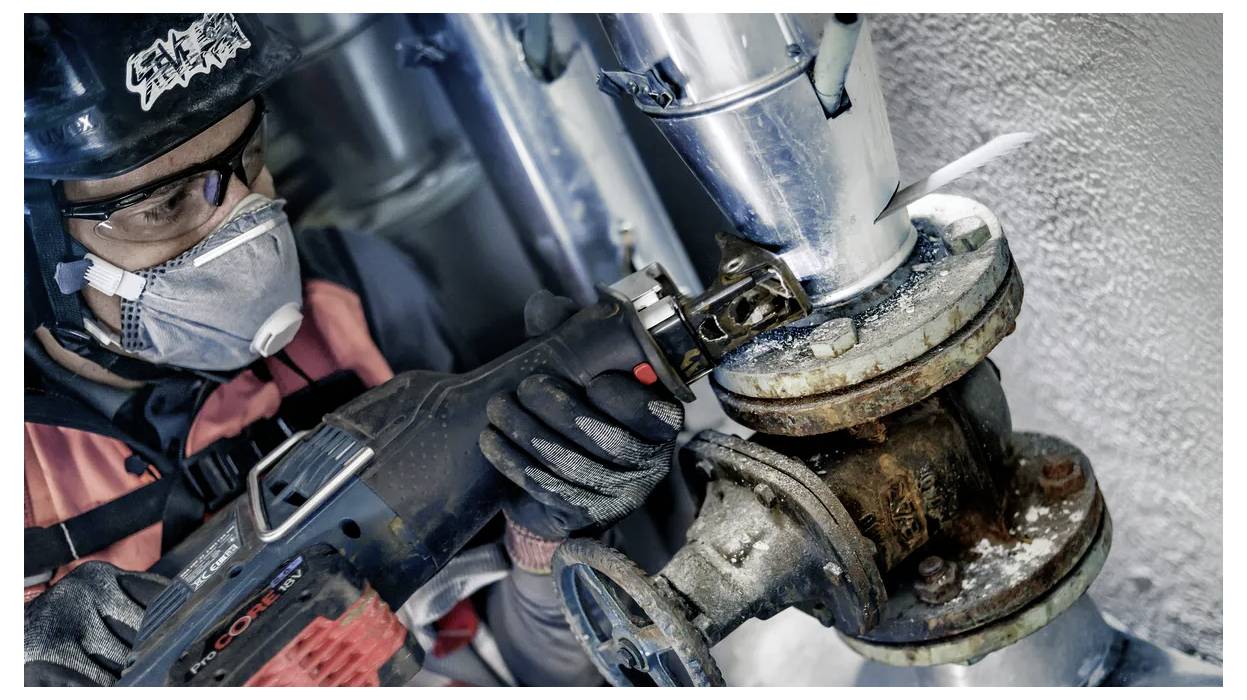 A worker wearing safety gear, including a helmet and mask, uses a power tool to cut through a rusted metal pipe in an industrial setting.