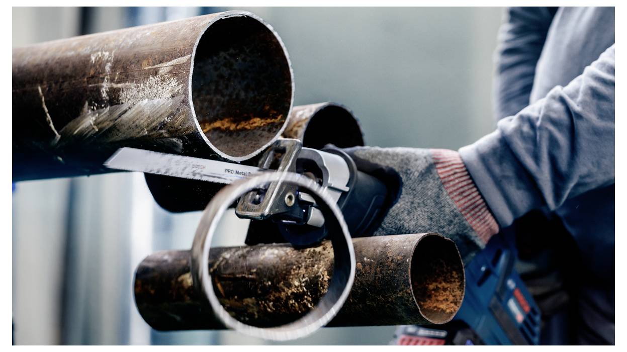 A person in protective gloves uses a handheld saw to cut through a large, rusted metal pipe, focusing on a precise cut.