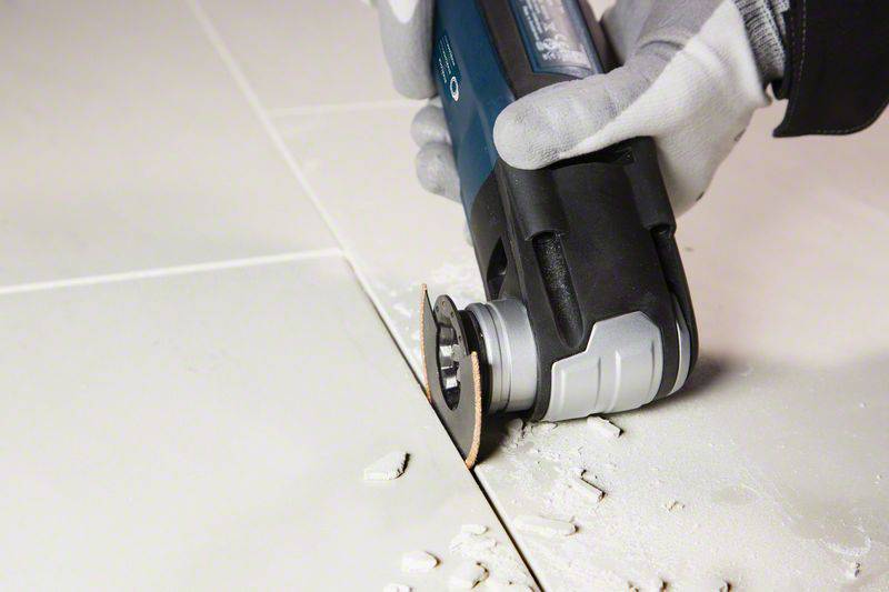 A person wearing gloves is using an electric multi-tool to cut tiles. Dust and fragments are visible.