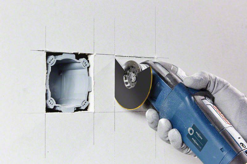 A hand holds an oscillating multi-tool, cutting an opening in a wall for a flush-mounted socket box.