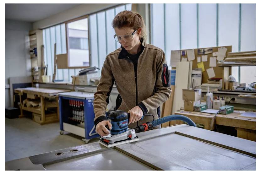 'Person sanding a large piece of wood with a power sander in a workshop. Shelves with tools and materials are in the background.'