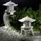 Two stone garden lanterns, one large and curved, the other small and simple, surrounded by green ferns and white pebbles.