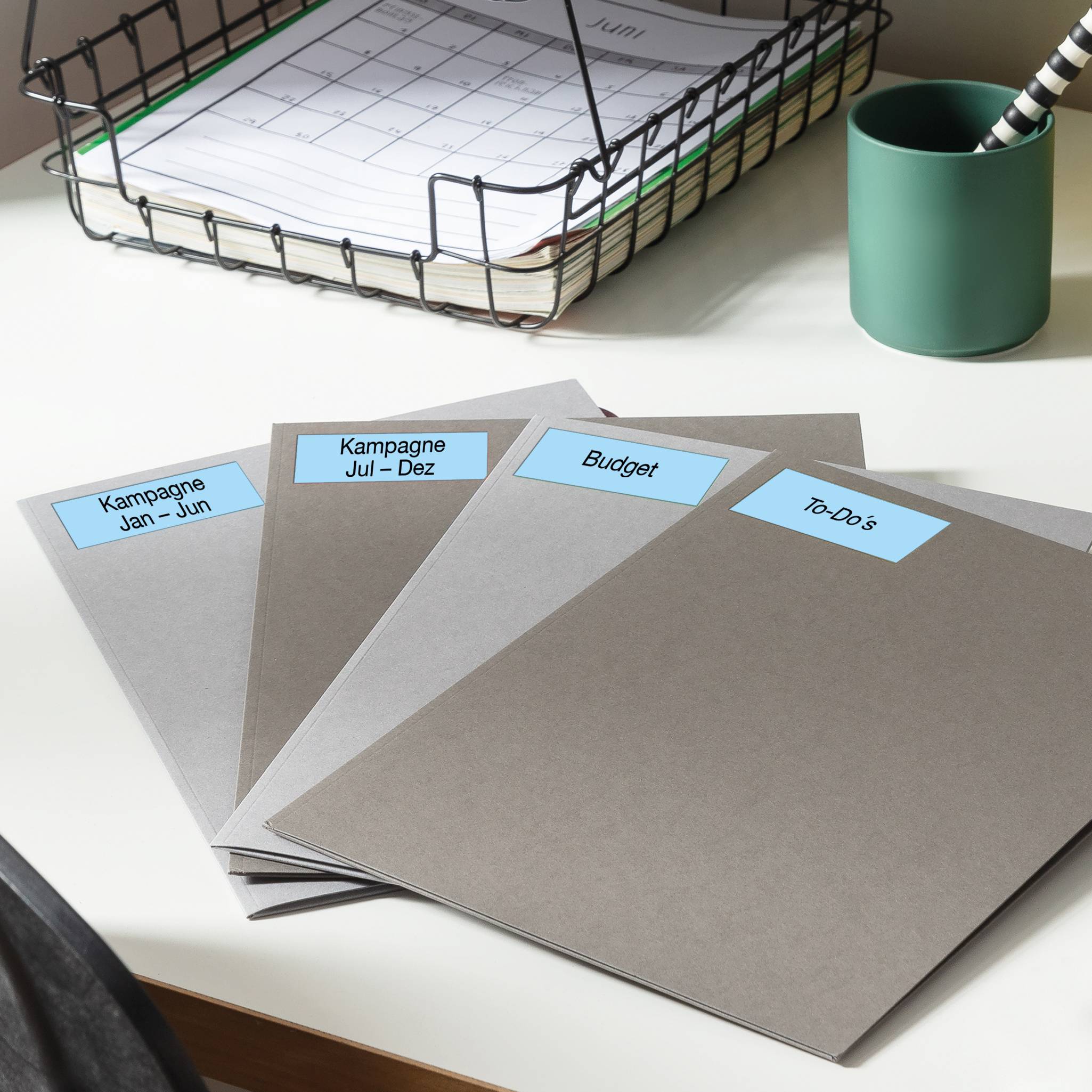 Pile of folders on a desk, labelled 'Campaign Jan - Jun', 'Campaign Jul - Dec', 'Budget', 'To-Do's'. A calendar is visible in the background.