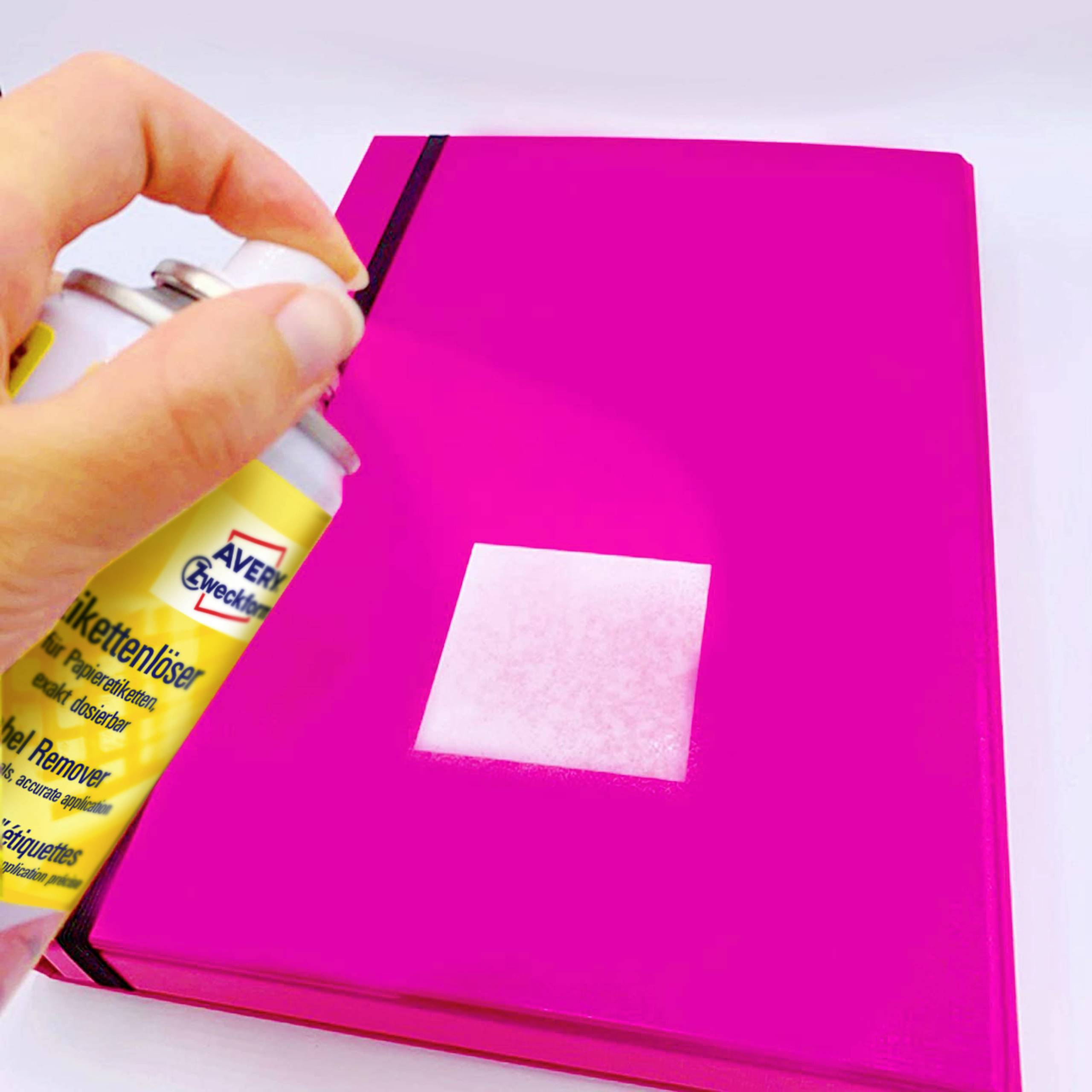 A hand sprays label remover onto a pink book, removing a rectangular label.