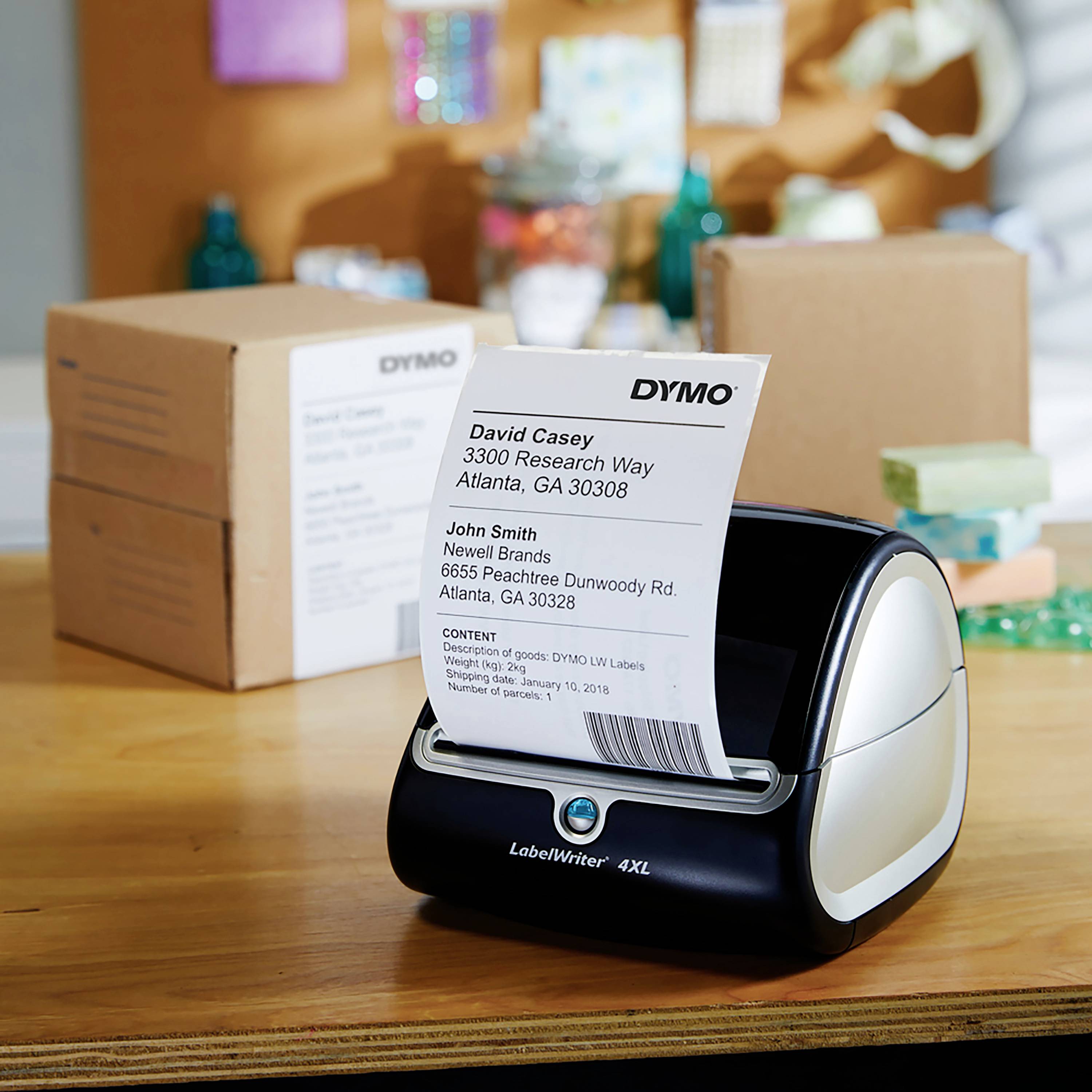 A label printer is printing an address label. Packaging boxes are visible in the background. Main focus is on the printing process.