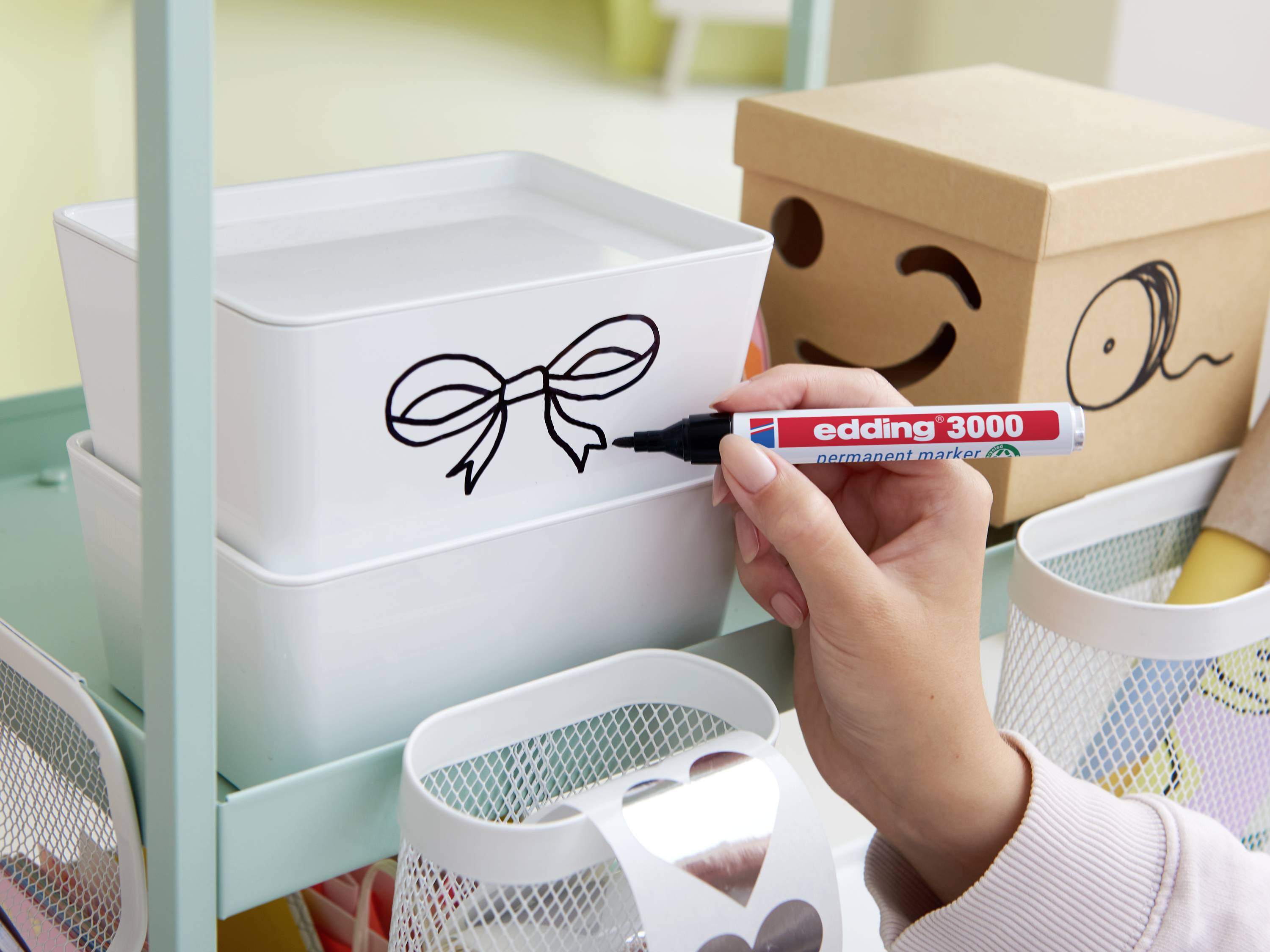 A hand draws a loop with a red marker on a white box. In the background are colourful boxes and a shelf.
