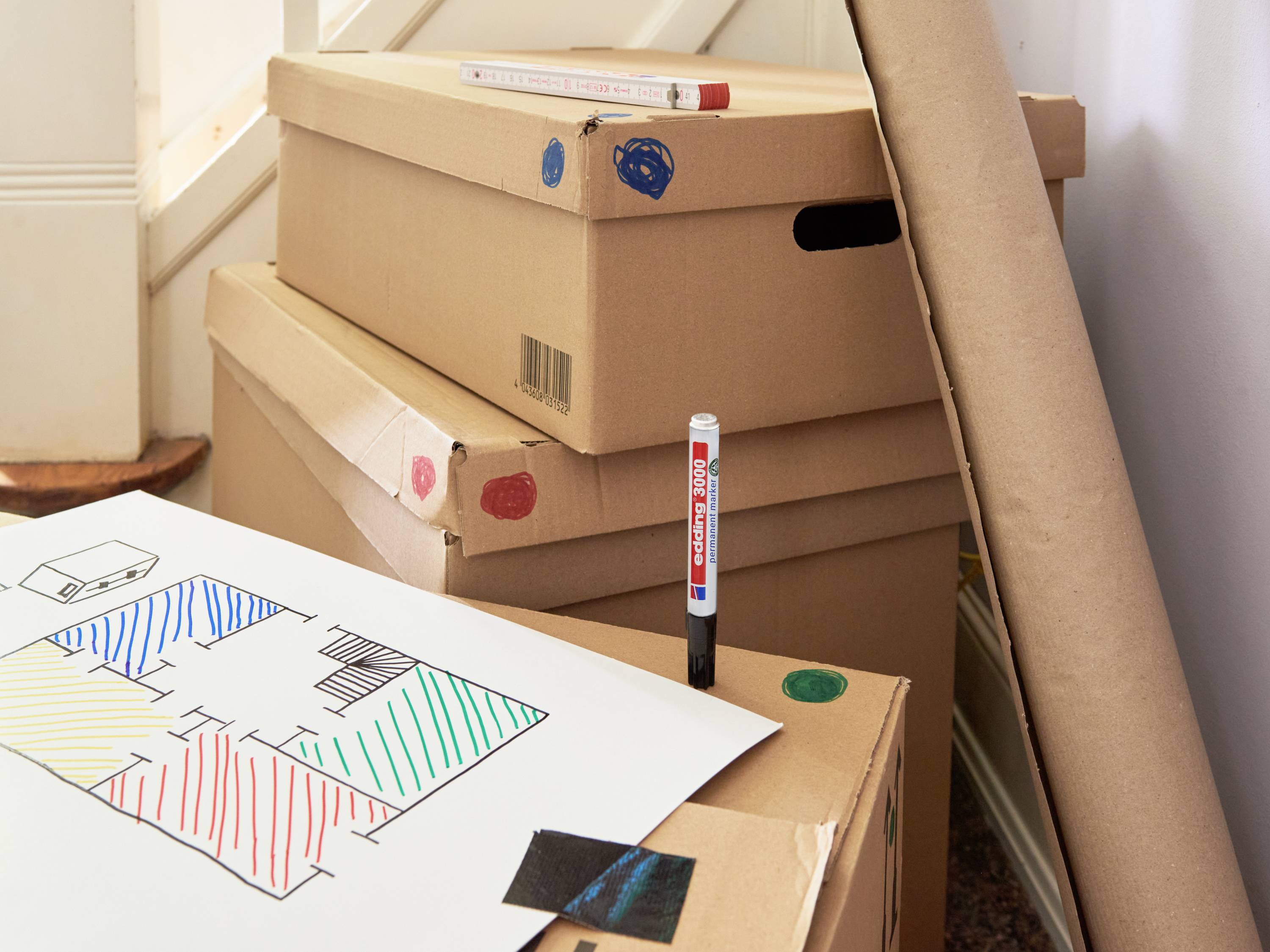 Cardboard boxes are stacked in a room with a staircase. A floor plan with coloured markings lies atop one of the boxes.