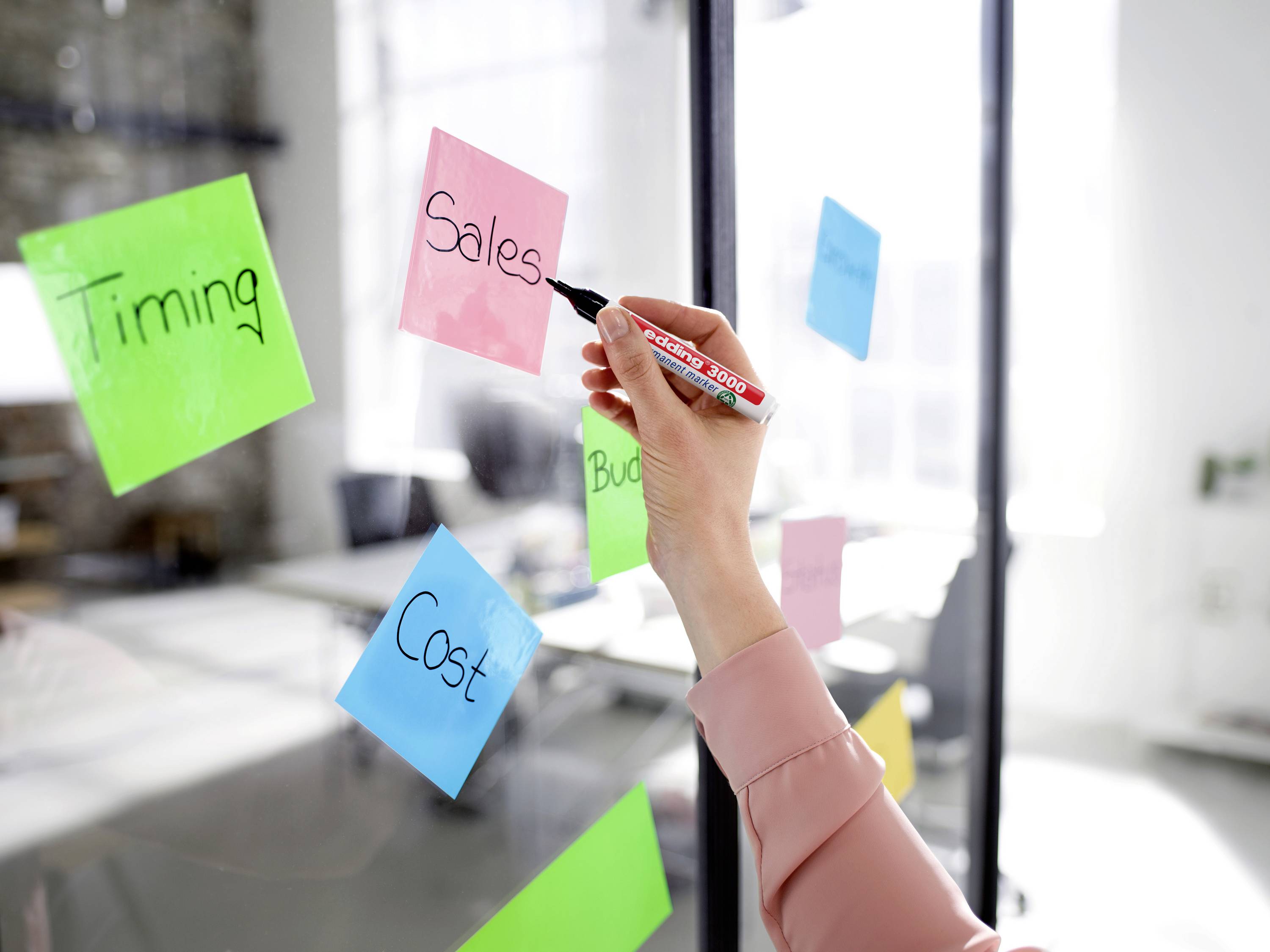 A person is writing with a marker on sticky notes attached to a glass wall. The notes display words like 'Sales' and 'Budget'. An office environment is visible in the background.