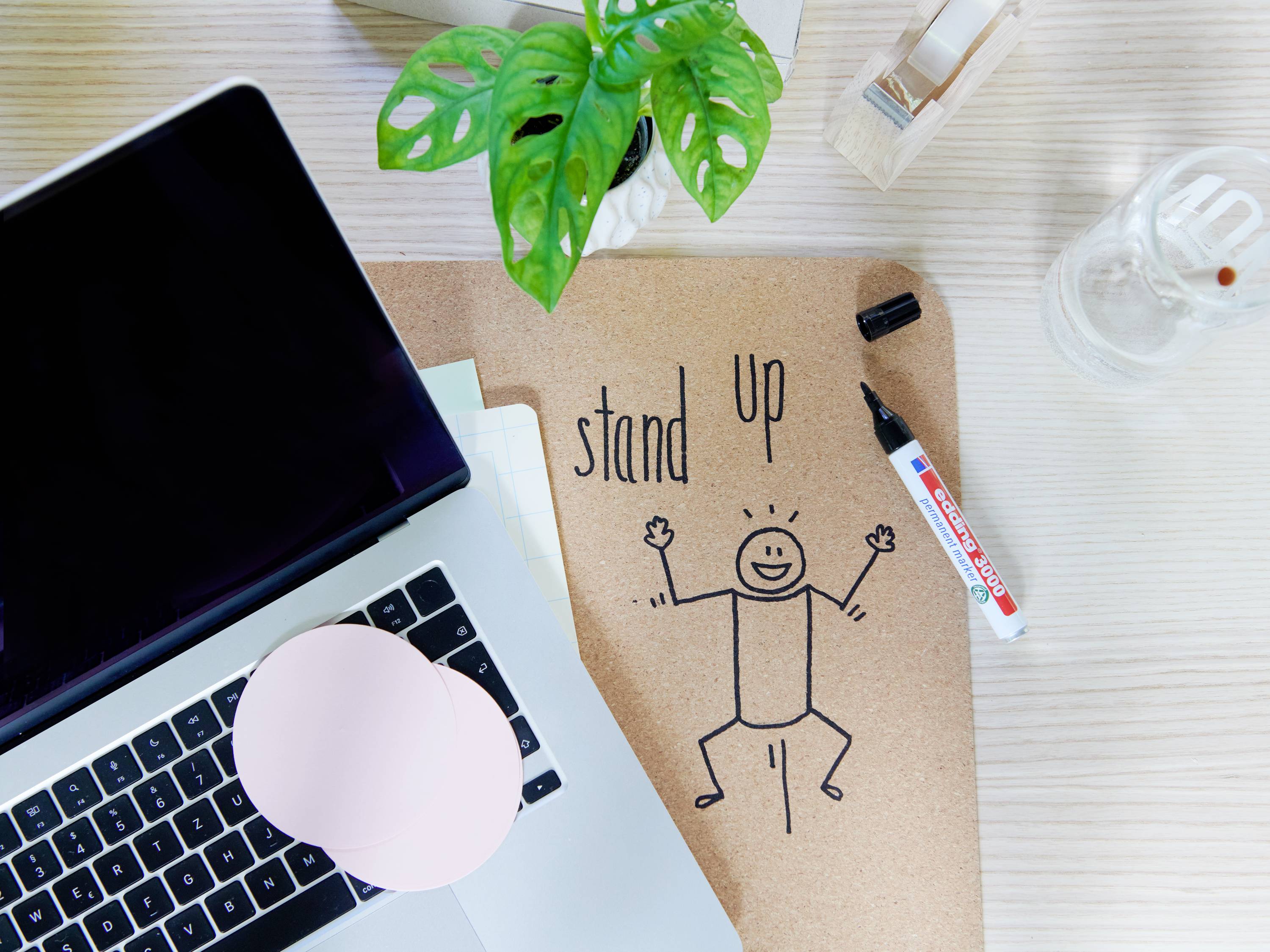 Laptop on a desk with a 'stand up' note and a drawn figure, alongside a pen and a plant, encourages movement in everyday life.