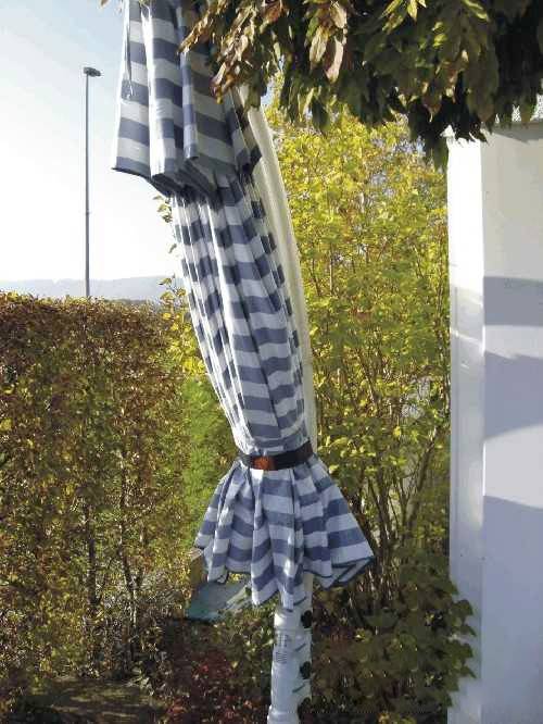 A blue and white striped parasol is closed and leaning against a white wall, surrounded by green bushes and trees.