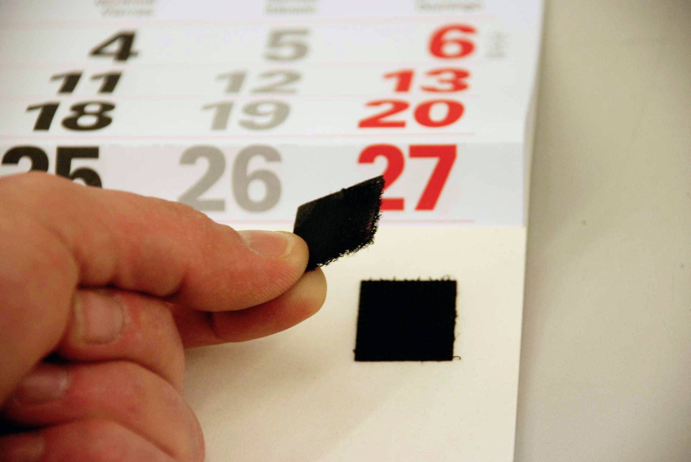 A hand removes a black adhesive label from a wall calendar, with the 27th marked in red.