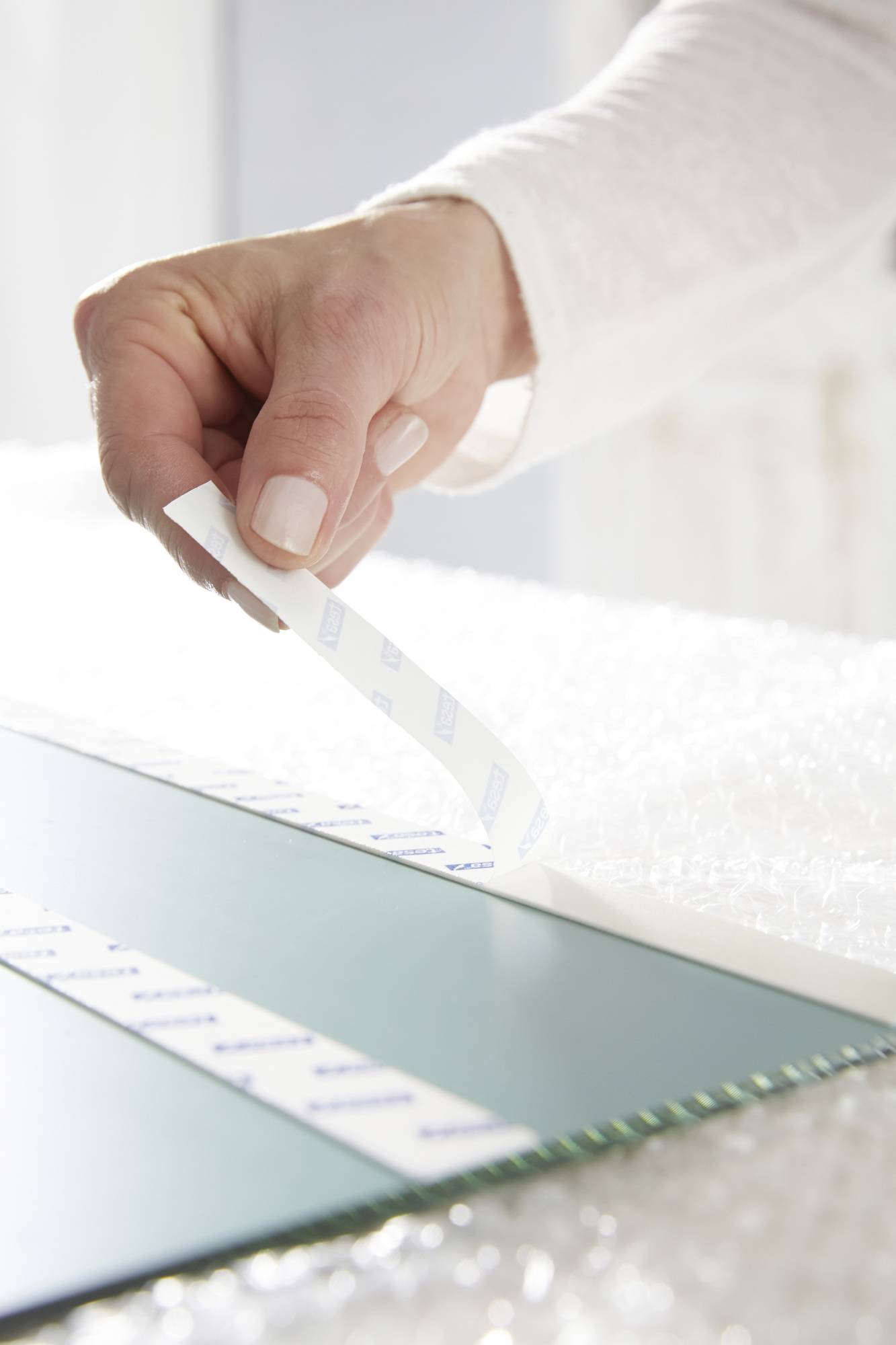 A hand carefully removes a piece of double-sided adhesive tape from the surface of a glass sitting on bubble wrap.