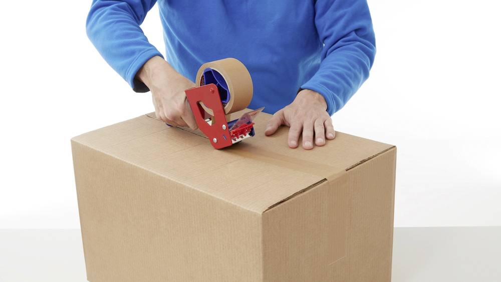 A person is packing a cardboard box with adhesive tape. The person is wearing a blue jumper and using a red parcel tape dispenser.