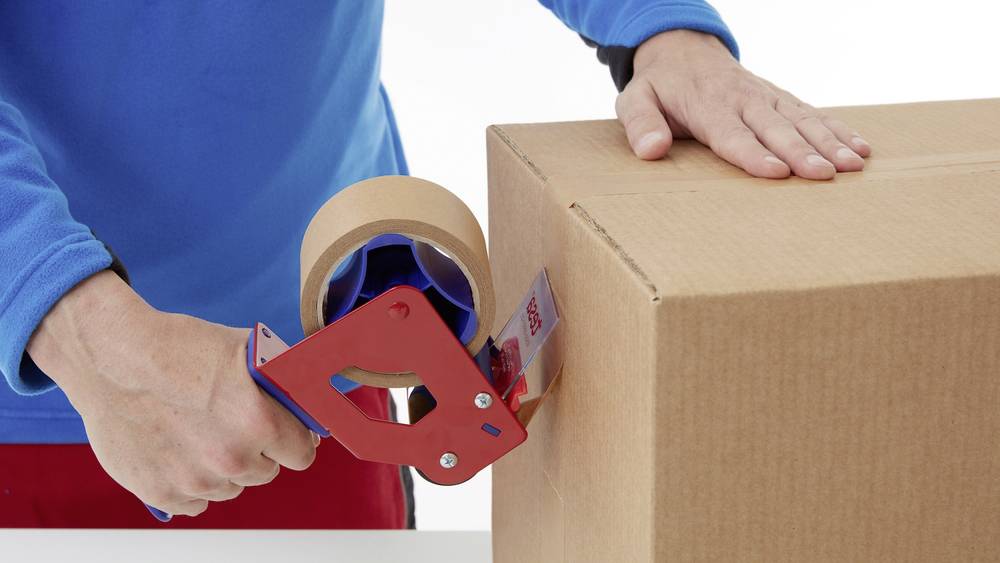 A person is sealing a cardboard box using a blue and red parcel tape dispenser on a white table.