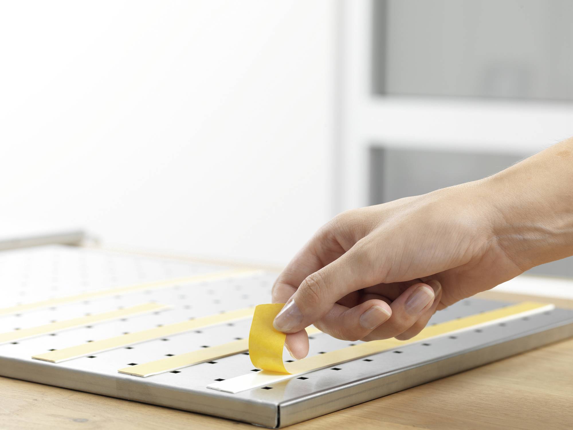 A hand lifts yellow longitudinal strips from a perforated metal surface. A window is blurred in the background.