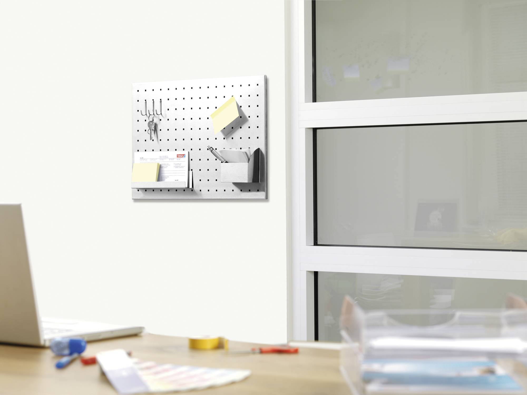 A desk scene with a laptop in the foreground and a perforated wall organiser with office supplies hanging on the wall in the background.