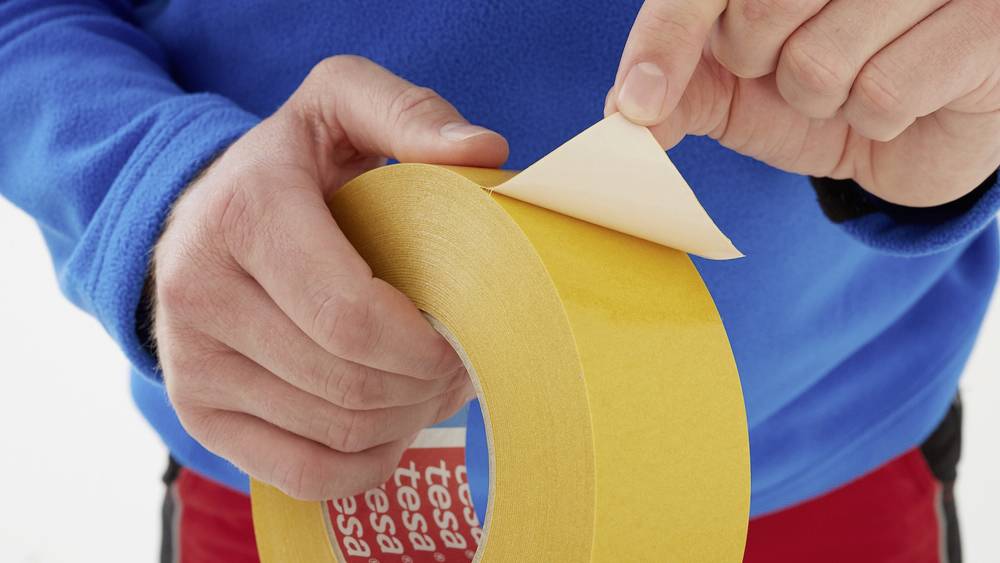 A person is peeling off the end of a yellow double-sided adhesive tape, ready for use.