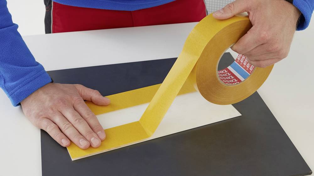 A person is sticking yellow adhesive tape onto a rectangular piece of paper on a table. Hands are visible as the tape is being unrolled.
